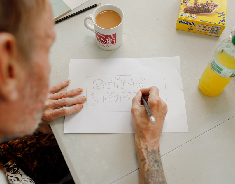 Person drawing the words 'Being Stronger' during a creative workshop, with a mug of tea and drink on the table