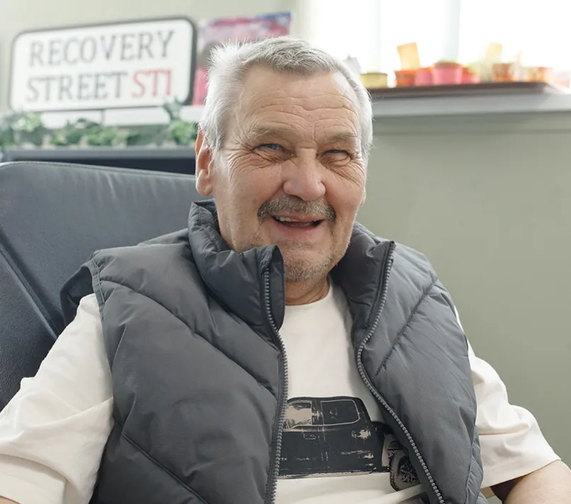 Smiling man sitting in a relaxed setting, with a sign that says 'Recovery Street ST1' in the background