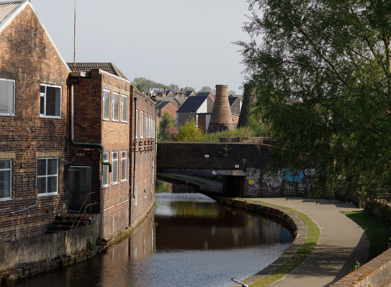 A canal towpath curving alongside sill water in Stoke-on-Trent, with traditional bottle kilns visible in the background among rooftops and trees