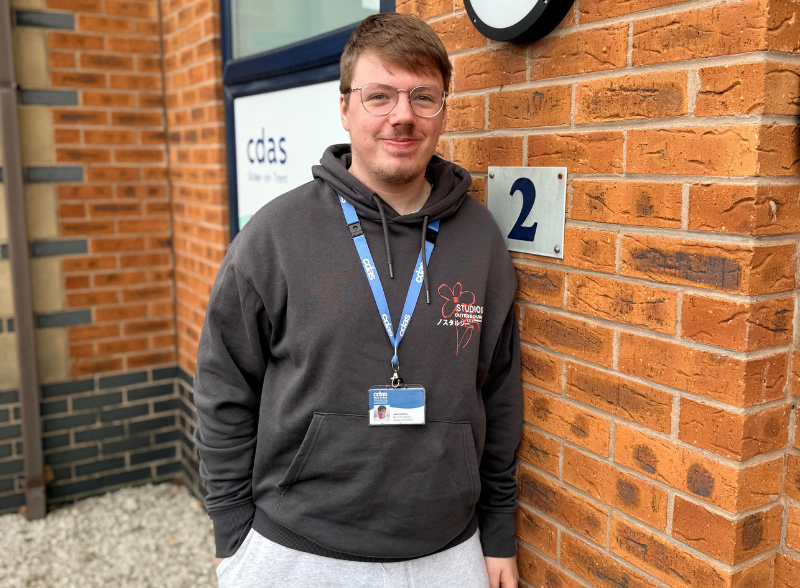 A photo of Josh, leaning against a brick wall with a silver number 2 plaque. He wears a grey hoodie, grey trousers and a CDAS lanyard and badge. Behind him is a window with the CDAS logo just over is right shoulder.