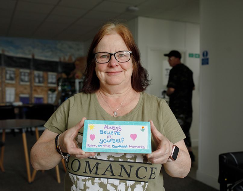 A smiling woman holds a homemade sign that reads 'Always believe in yourself even in the darkest moments'