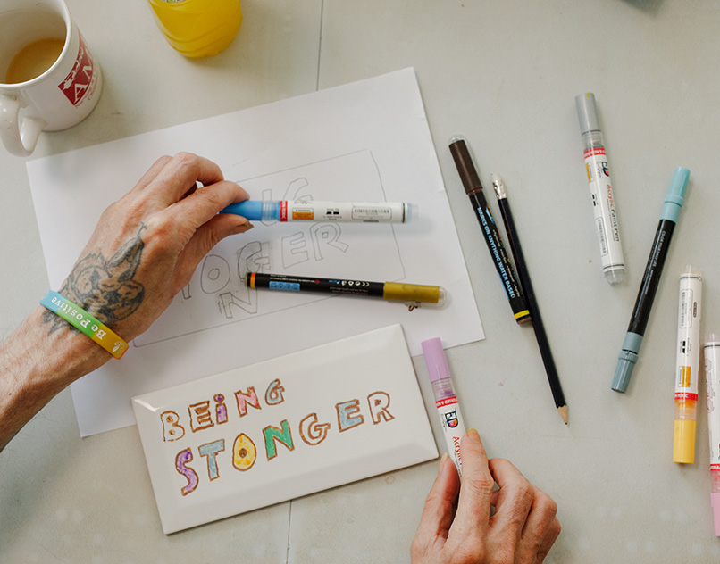Hands creating artwork with the words 'Being Stronger' during a creative session, representing support for friends and family